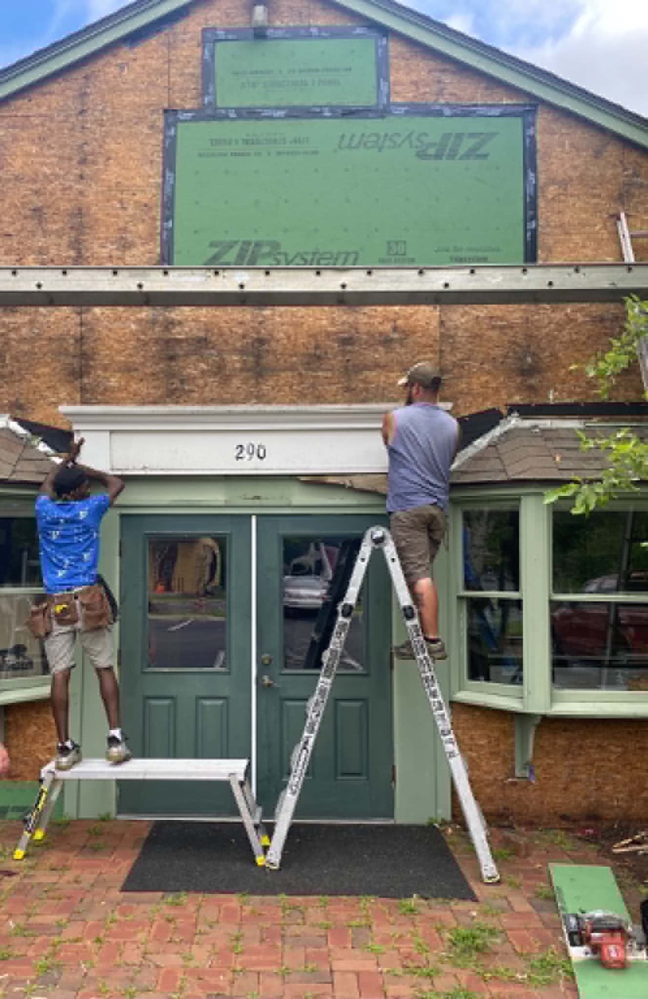Two carpenters on ladders reworking the entryway and trim of a green storefront