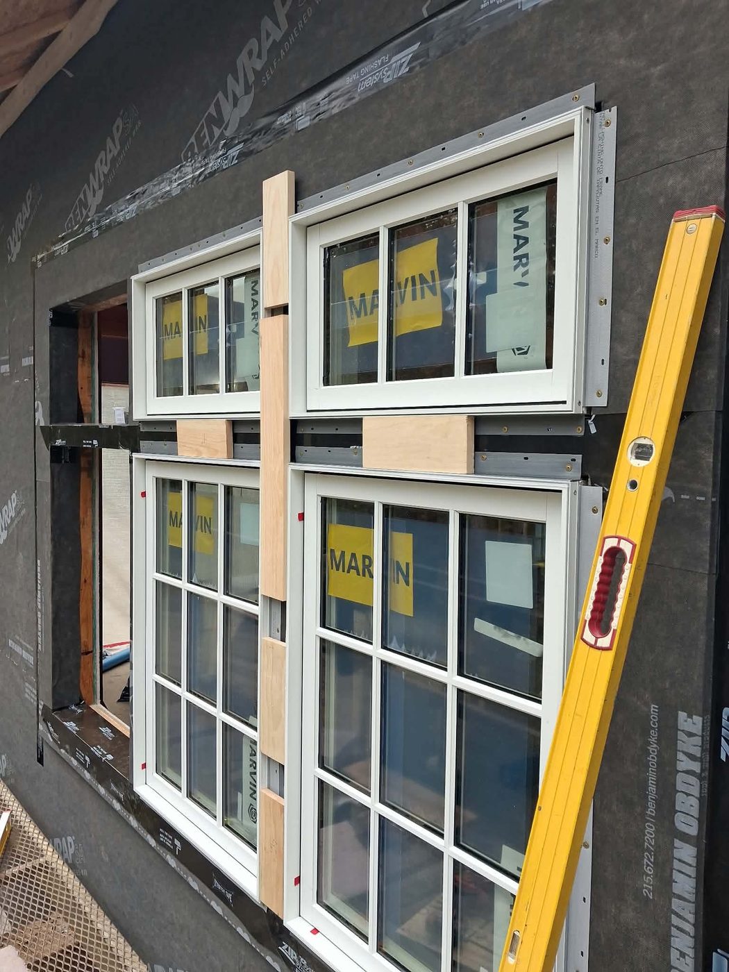 New white windows being set in their openings, with wood spacer blocks between the units and a yellow level against the frame