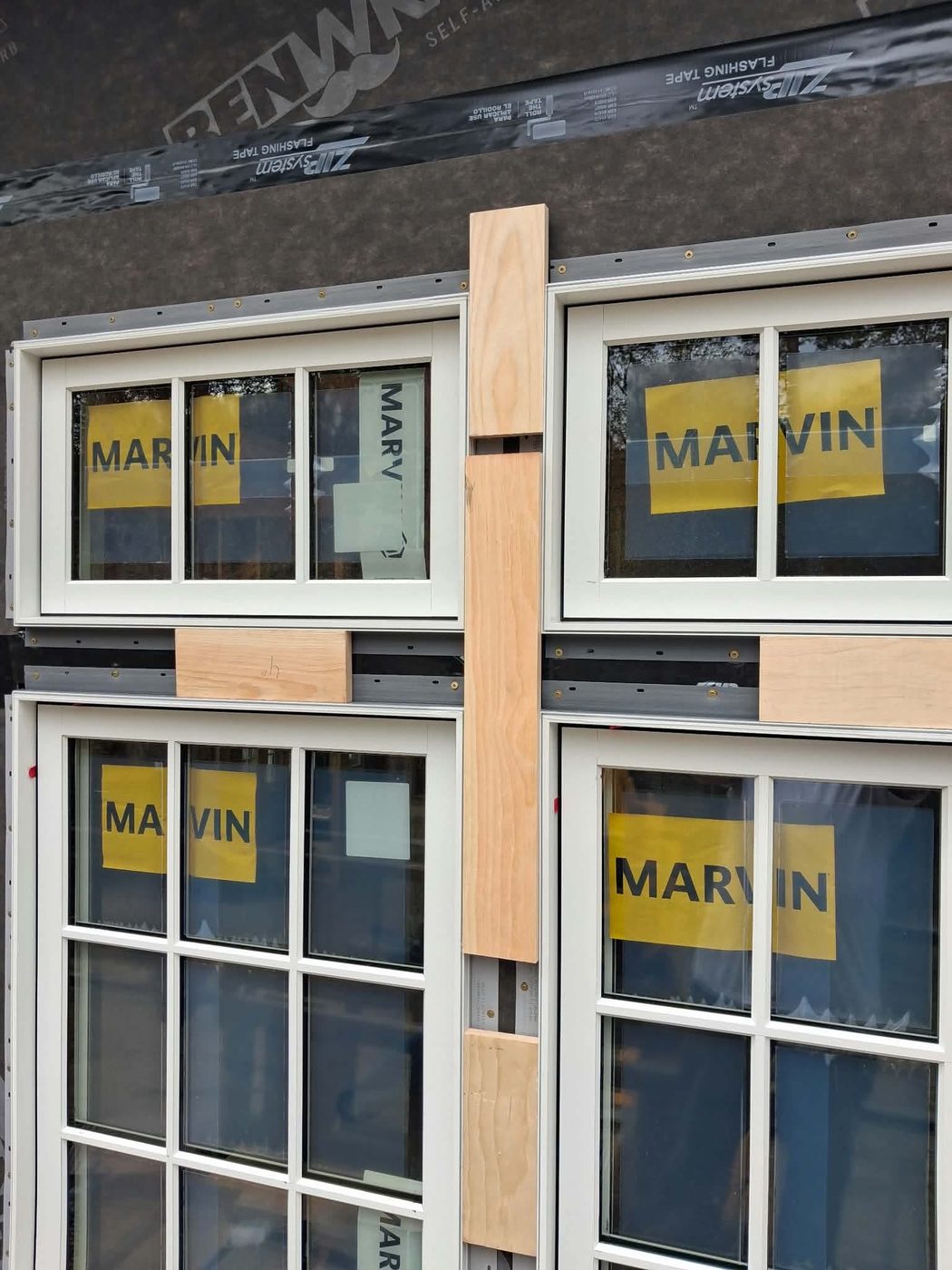 Close-up of paired windows with factory stickers still on the glass, wood blocking between the units and flashing tape across the head